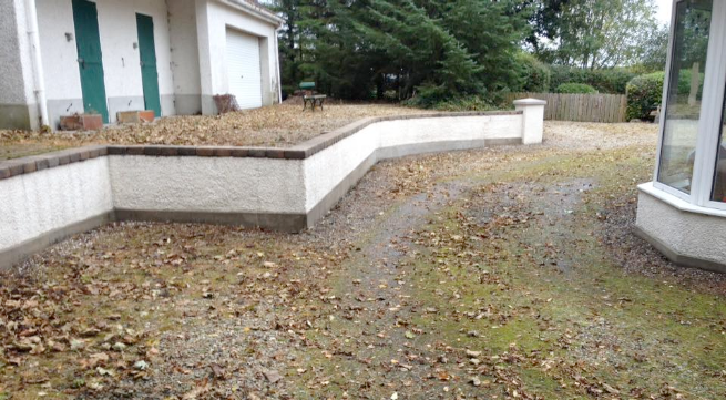 Overgrown driveway with moss and leaf debris beside a rendered garden wall